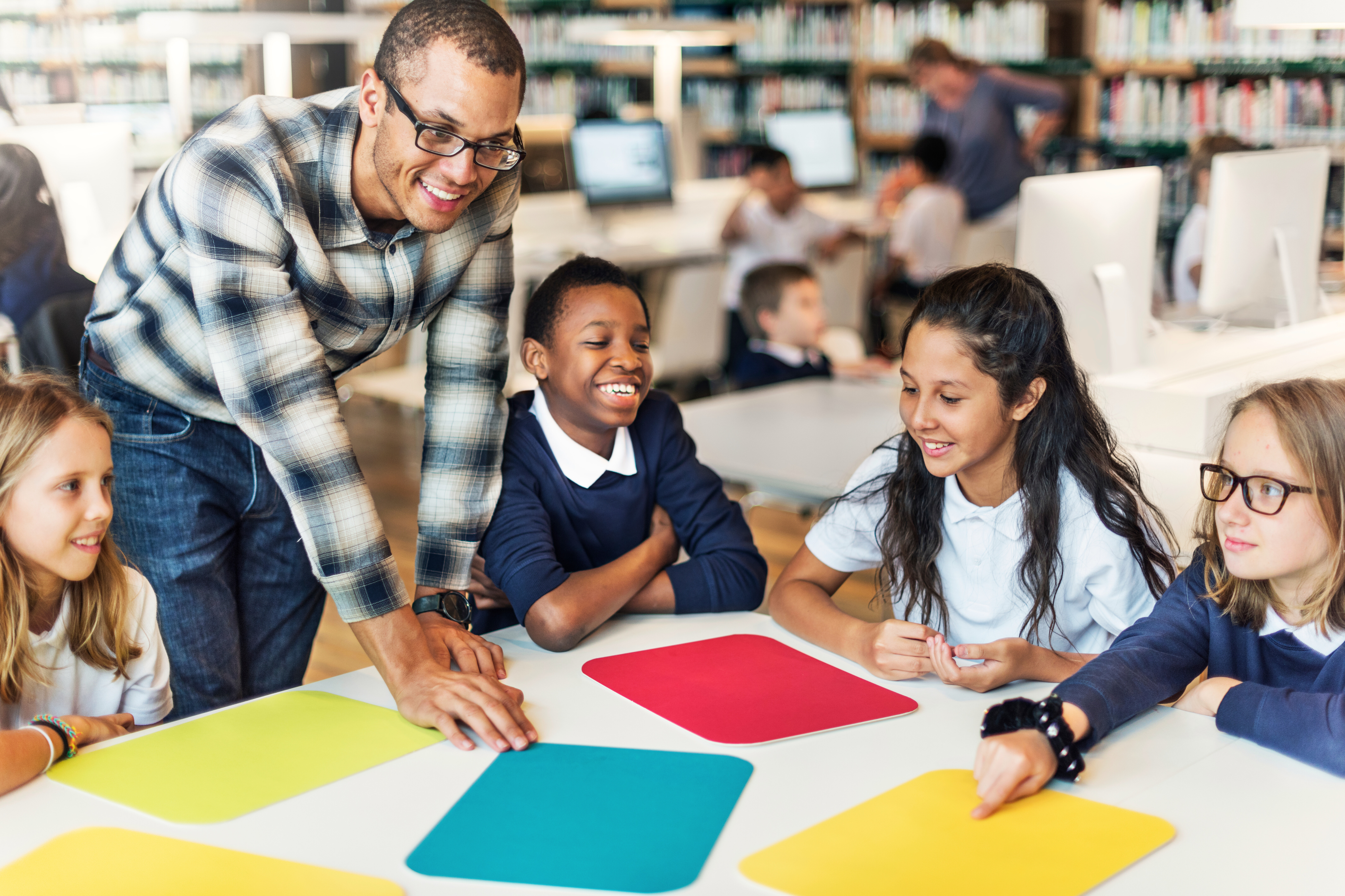 K-12 ERP Solutions can help streamline processes to free up time for what's important - Helping your students! Pictured is an adult male teacher working with a group of smiling kids with coloured duotangs.