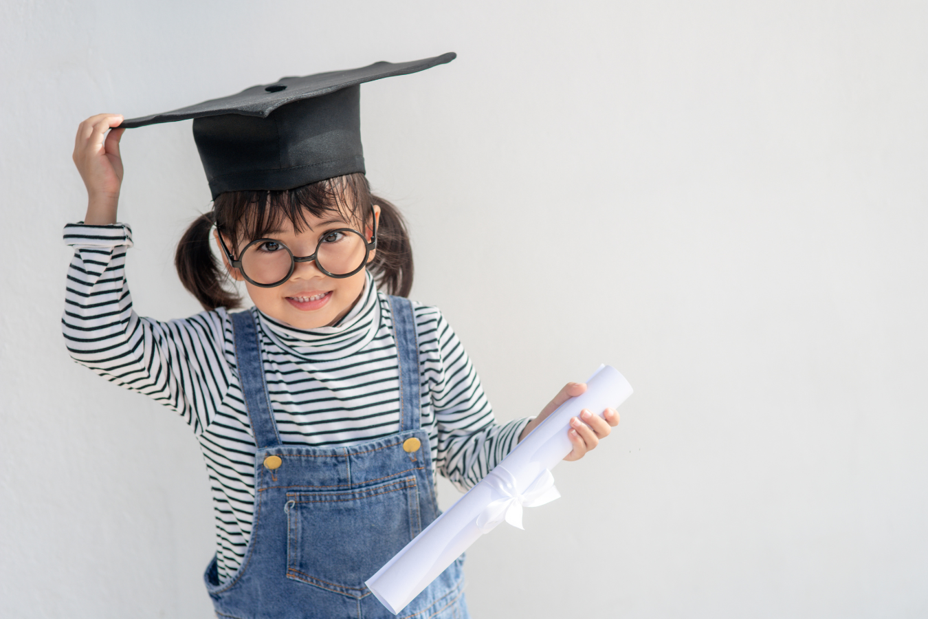 A young female kindergarten smiles as she touches the graduation cap on her head and holds a rolled up diploma in her other hand. Sparkrock 365 is an ERP for K12 and education that can be maximized to optimize school operations.