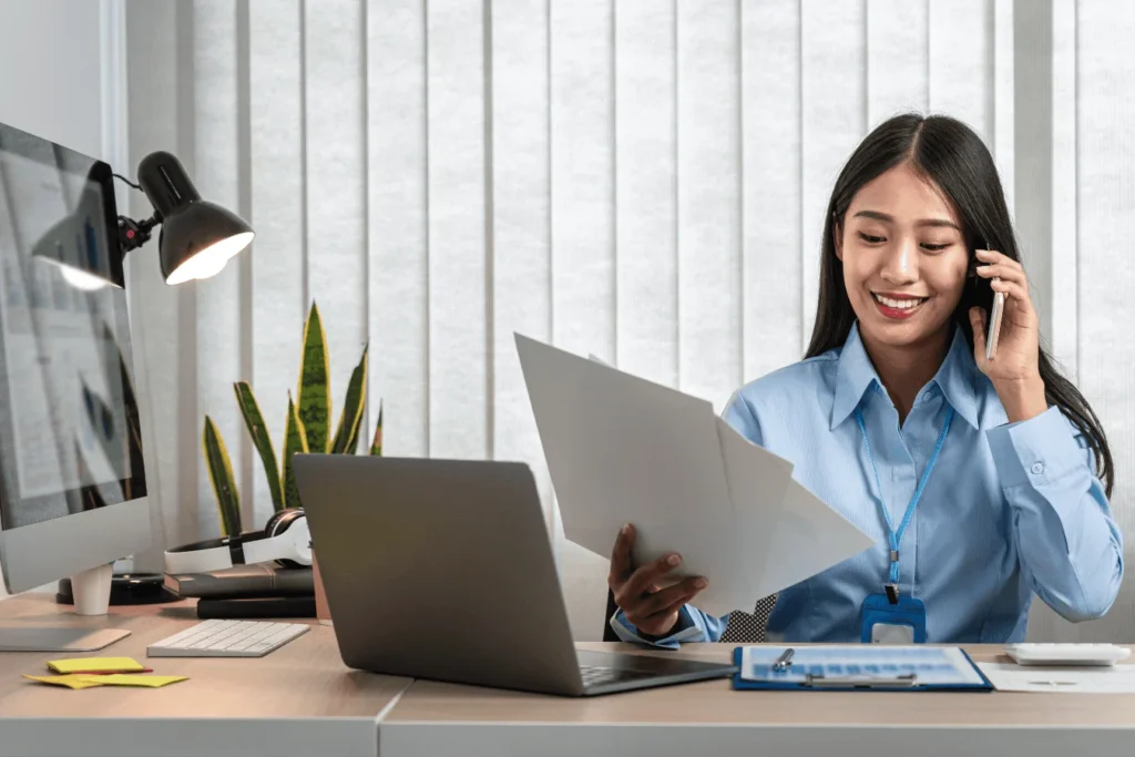 Female IT staff member smiling while on the phone, reviewing documents at her desk with a computer and laptop in a school office setting.
