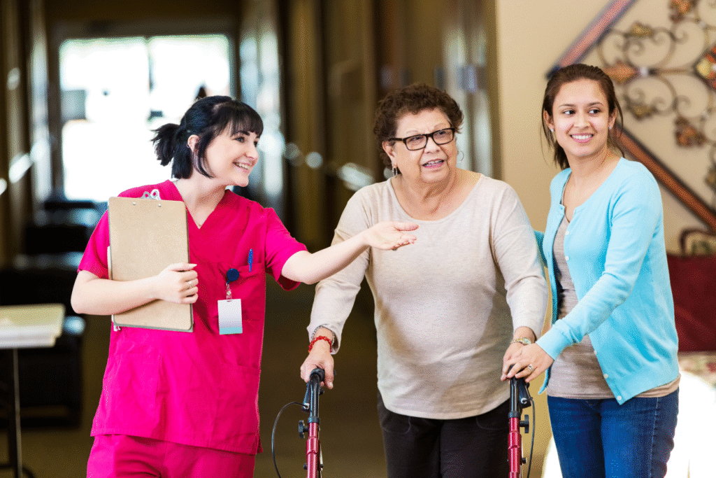 A female nurse in neon pink scrubs points an older woman and her daughter in the right direction in an assisted living facility's hallway.