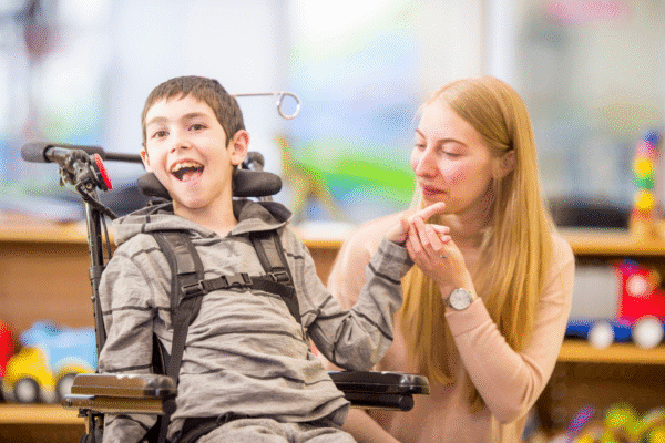A smiling young boy in a wheelchair holds hands with a supportive caregiver in a bright, welcoming environment, illustrating compassion and connection in disability care.