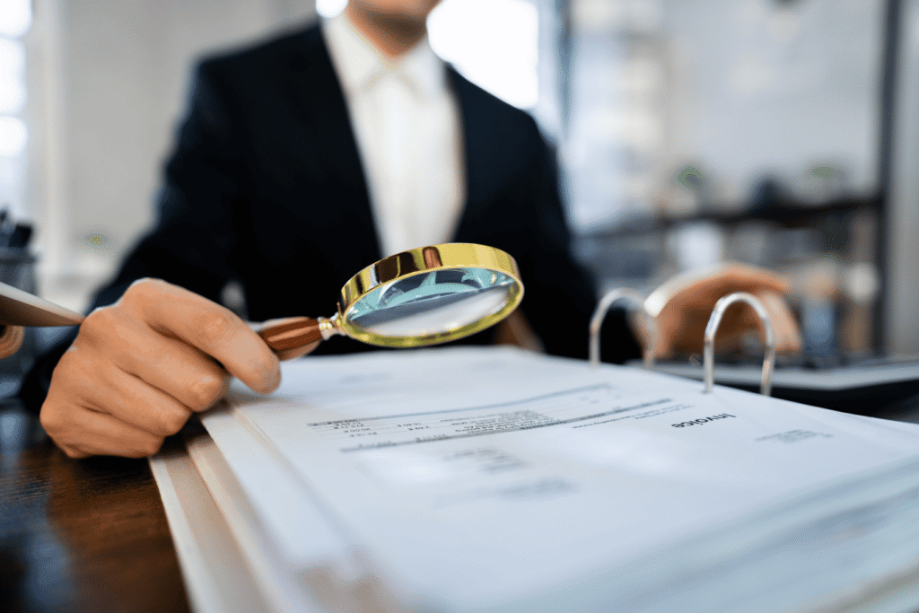 A person wearing professional business attire is holding a magnifying glass, closely examining an invoice. The invoice is partially visible, featuring rows of text and numbers. The individual appears focused, symbolizing careful financial analysis or auditing. The background is minimal, keeping the focus on the person and the invoice.