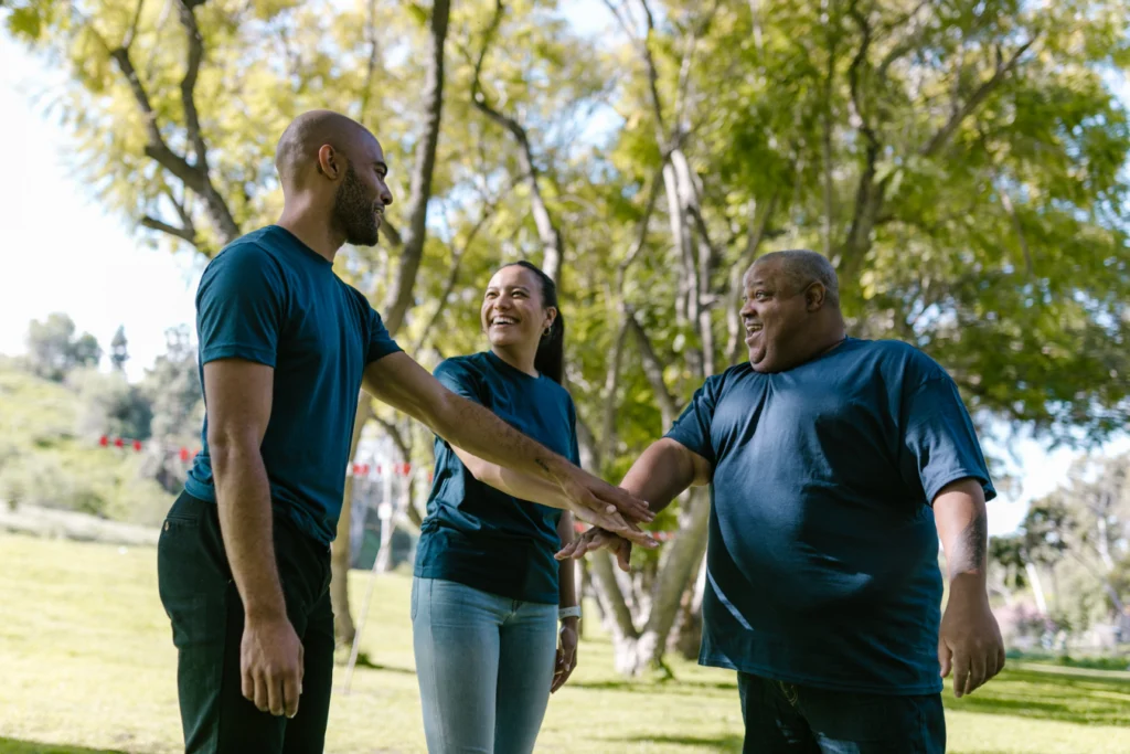Three people wearing matching navy t-shirts stand in a park, smiling and placing their hands together in a team huddle. The background features green grass, trees, and sunlight filtering through the leaves.