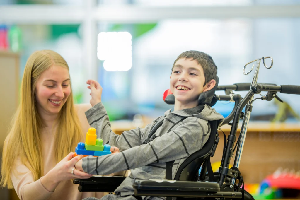 Smiling caregiver assisting a young boy in a wheelchair as he plays with colorful building blocks, symbolizing empowerment and inclusion for individuals with disabilities.