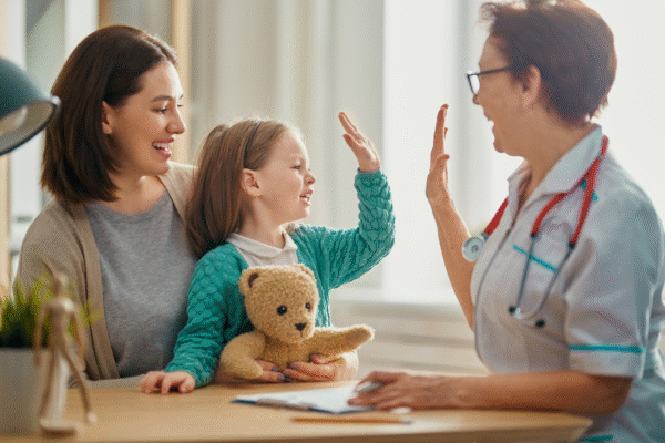 Smiling young girl holding a teddy bear gives a high-five to a doctor in a white coat, while sitting on her mother’s lap during a cheerful medical appointment.