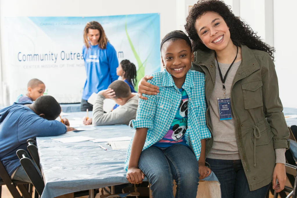 A smiling youth counselor stands with her arm around a young girl at a community outreach program. In the background, several children work on activities at a table while another mentor offers guidance. The atmosphere is positive and supportive.