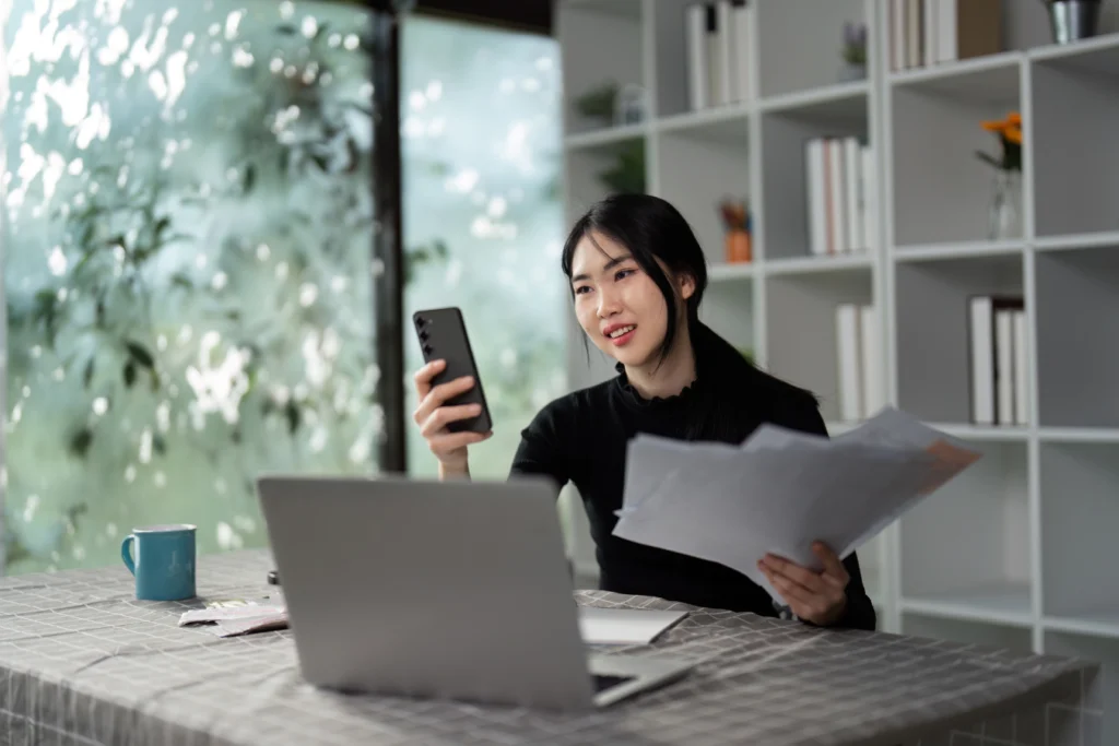 Young professional holding receipts and checking expense management app on her phone while working on a laptop at home office.