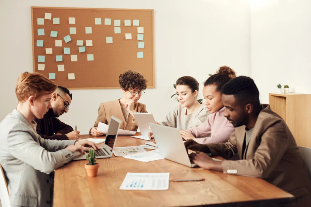 Six colleagues sit around a wooden conference table, working together with laptops, tablets, and documents. A corkboard with colorful sticky notes is visible on the wall in the background, and small potted plants decorate the table, creating a bright and collaborative office atmosphere.