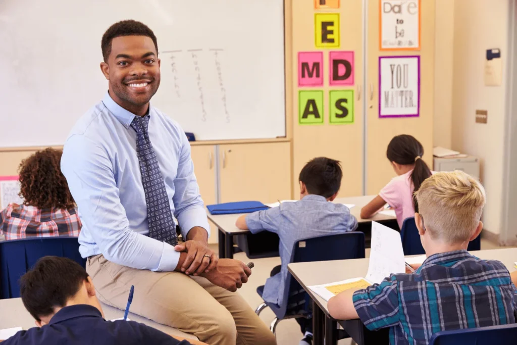 Male teacher smiling at the camera while students work at their desks in a bright, connected classroom setting.