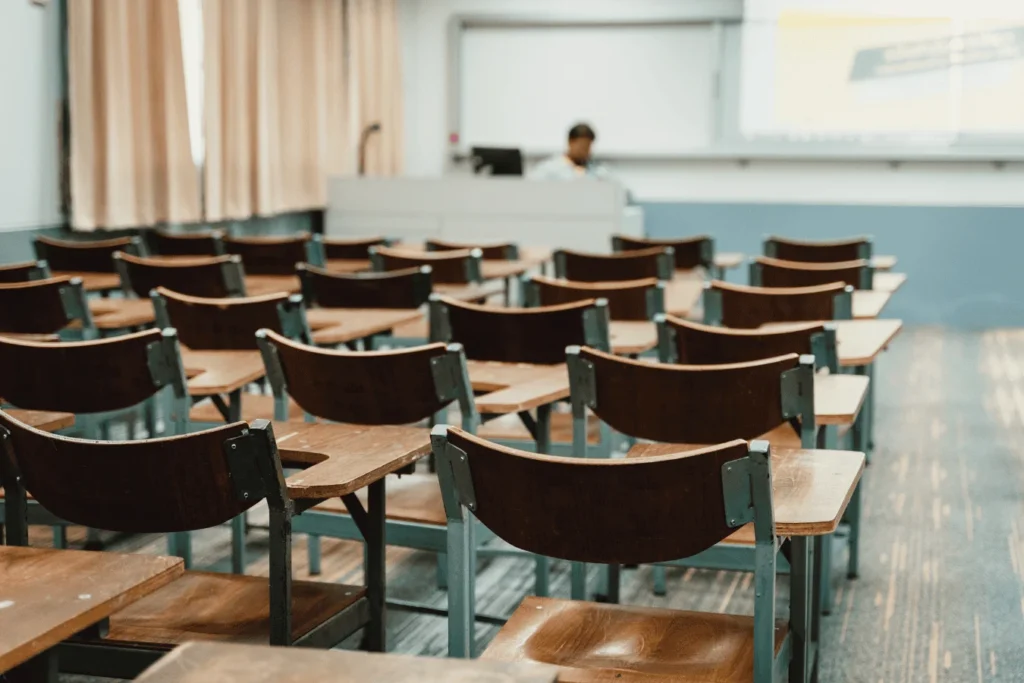 Rows of empty wooden desks in a classroom with a teacher at the front, emphasizing absenteeism and student tracking issues.