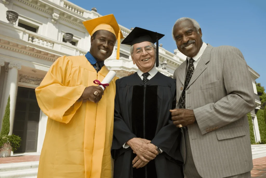 A graduate in a yellow cap and gown stands outside a university building, smiling alongside a faculty member in academic regalia and a family member in a suit.