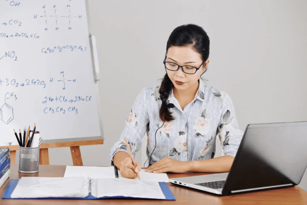A teacher sits at a desk with a laptop and open binder, taking notes while reviewing data. A whiteboard with chemical equations is in the background.