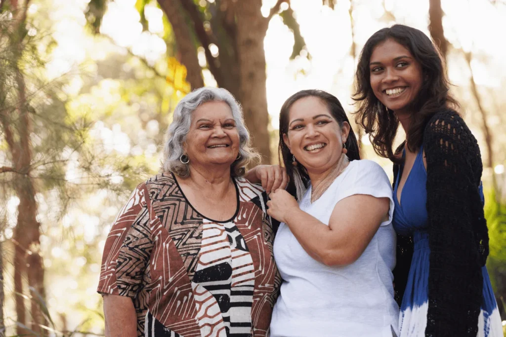 Three Indigenous women from different generations standing outdoors, smiling and enjoying time together in their community.